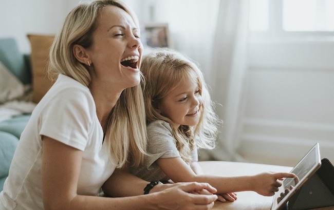 A woman and her daughter are sitting together on the floor; the woman is laughing, and her daughter is using a tablet and smiling.