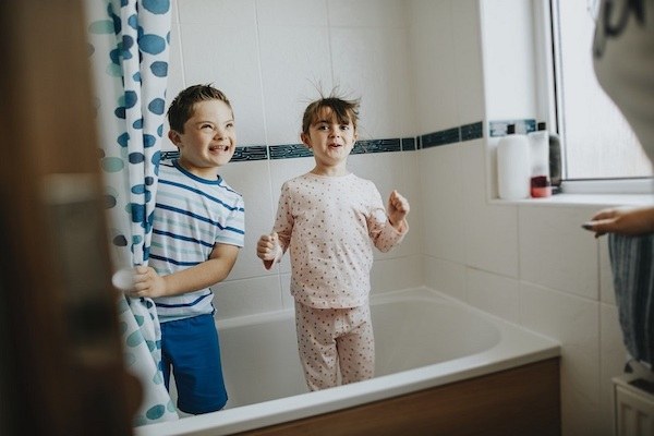 Two children, a boy and a girl, are standing up in a bathtub with their pajamas on, and are smiling.