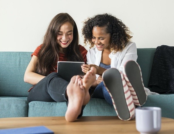 Two teens are sitting on a couch, with their feet on a coffee table, and looking at a tablet; they are smiling.