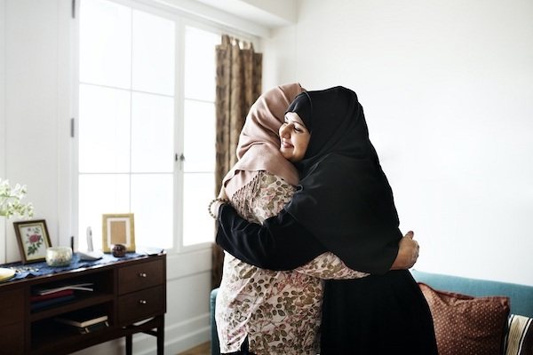 Two women are standing up and hugging in a living room; one has a big smile on her face.