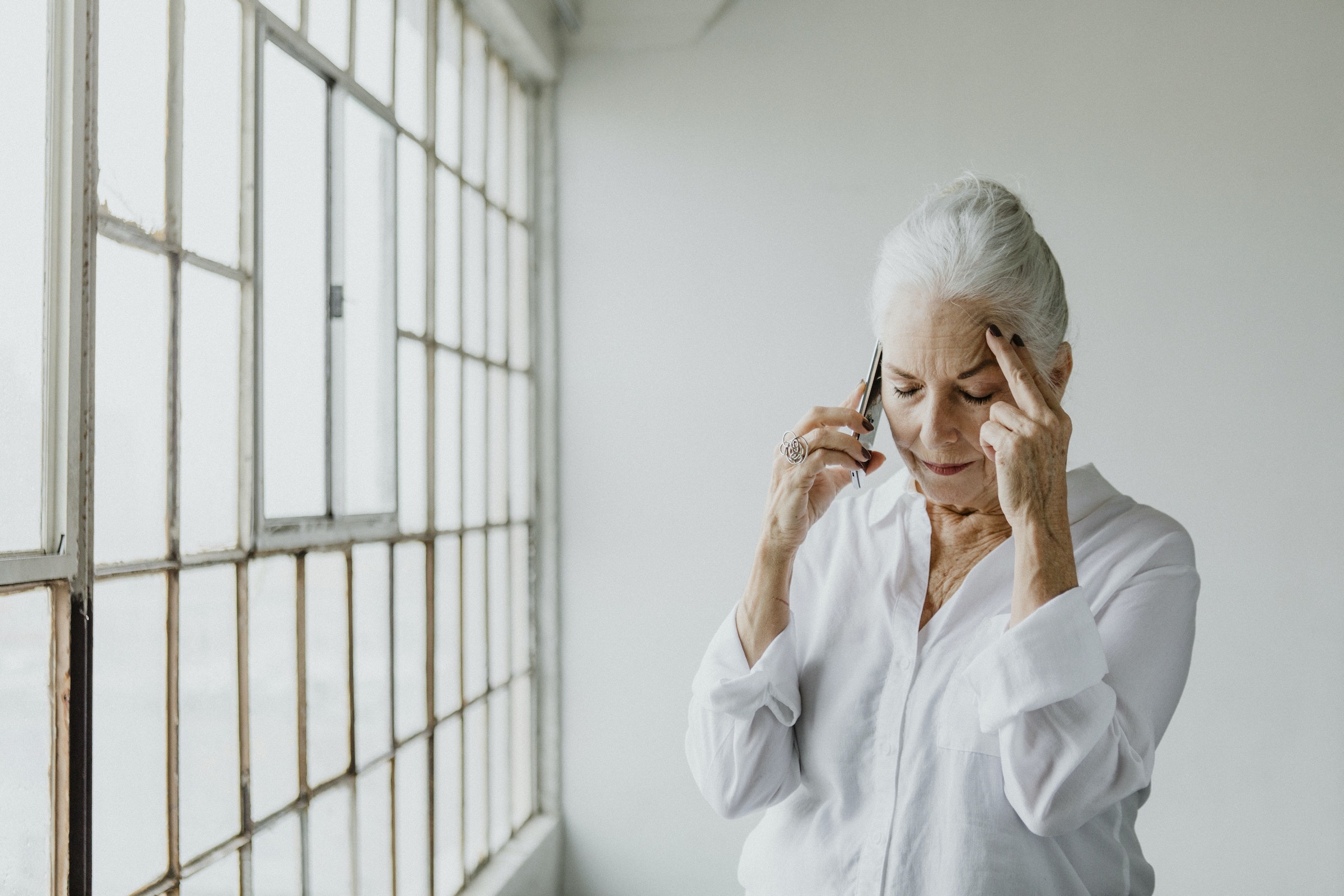 frowning woman with white hair places fingers on her forehead in frustration while holding a cell phone to her ear